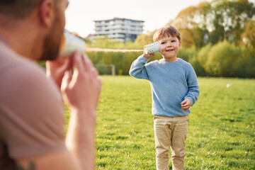 Wall Mural - Using paper cup telephone. Happy father with son are having fun on the field at summertime