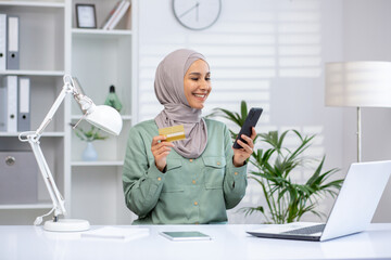 Smiling woman in hijab using credit card and phone for online shopping while working in a home office setup.