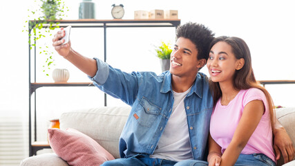 Wall Mural - A teen couple sits on a couch in a living room and takes a selfie with a smartphone. The guy is wearing a denim shirt and jeans, while the girl is wearing a pink shirt and jeans. They appear happy