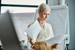 © LIGHTFIELD STUDIOS - A middle-aged businesswoman with short hair holds a piece of paper in front of a computer screen.
