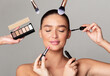 © Prostock-studio - A woman with eyes closed is having various makeup products applied on her face by multiple hands around her. She holds a serene expression as brushes and palettes are used to enhance her makeup look.