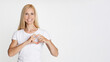 © Prostock-studio - A young woman with blonde hair is standing in front of a white background. She is wearing a white t-shirt and smiling. She is using her hands to make a heart shape in front of her chest.