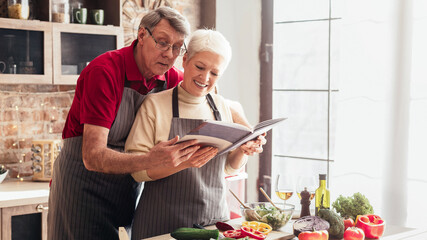 Wall Mural - An older couple wearing aprons is reading cooking book in their kitchen. They are smiling and appear to be enjoying themselves while preparing a meal together