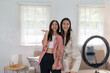 © amnaj - Two smiling women recording a video together indoors using a ring light and smartphone, with a cozy and bright room background.