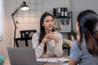 © amnaj - Two women having a professional conversation in a modern office setting, one taking notes on a laptop while the other listens attentively.