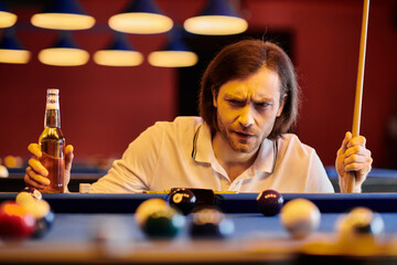 a man in casual attire focuses intently on the billiard table, holding a cue stick and a beer bottle