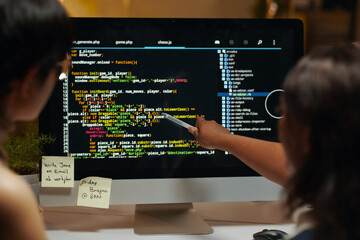 Developers pointing at programming code on computer screen and discussing, view from behind