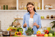 © Prostock-studio - A woman smiles while mixing a salad in her kitchen, surrounded by fresh vegetables and a pot on the counter.