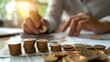 © Naret - Close-up of a person's hand calculating and stacking coins, representing finance, savings, and investment concepts in a bright setting.
