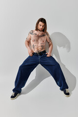 A man with a chest tattoo poses in front of a white wall.