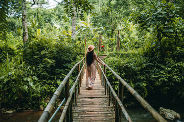 the young woman in a dress and hat is walking on the bridge, looking around in a tropical forest. le