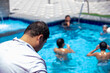 © Viga - A man in a pool, watching others swim. The background shows a lively poolside scene on a sunny day