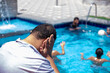 © Viga - A man in a pool covering his ears, possibly due to water inside them. The background shows a lively poolside scene under the bright sun