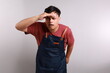© ArvStd - Portrait of curious asian man in apron holding hand above eyes and peering into distance, looking far away, expecting and searching someone on horizon. studio shot isolated