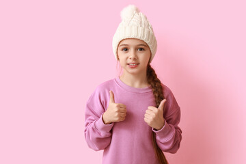 Wall Mural - Cute little girl in warm hat showing thumbs-up on blue background