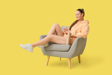 Canvas Print - Young woman with potato chips using mobile phone in armchair on yellow background