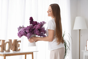 Canvas Print - Young woman putting vase of lilacs on table at home