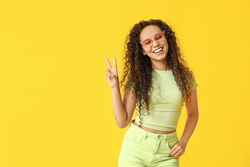 Wall Mural - Happy young African-American woman in sunglasses showing victory gesture on yellow background