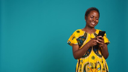 Wall Mural - Modern young woman browses online websites on her phone, standing against blue background. Cheerful satisfied girl checks her texts on social media, using a smartphone in studio. Camera B.