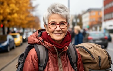 Wall Mural - A woman wearing a red coat and glasses is smiling and holding a backpack. She is standing on a street with cars and other people around her