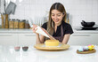 © chadchai - Young woman in an apron using a piping bag, pipe the icing onto the cake, creating a decorative pattern on the top of a round, light-colored cake that's placed on a wooden turntable or cake stand.