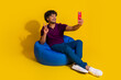 © deagreez - Full size photo of nice young man selfie photo v-sign sit bean bag device wear trendy violet outfit isolated on yellow color background