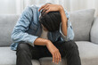 © KMPZZZ - Sad upset, frustrated alone asian young man headache thinking about finance problem, difficulty, feeling failure and exhausted, suffering from loneliness sitting on couch at home. Grief sorrow, bad.