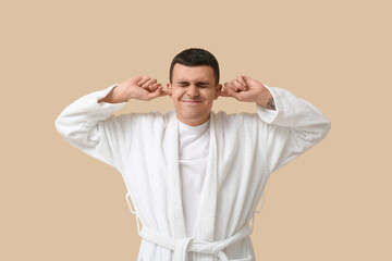 Wall Mural - Portrait of handsome young man in bathrobe suffering from loud noise on beige background