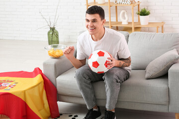 Wall Mural - Young man with soccer ball and flag of Spain watching football on sofa at home