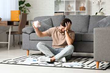 Wall Mural - Stressed young man in debt with piggy bank sitting on floor at home