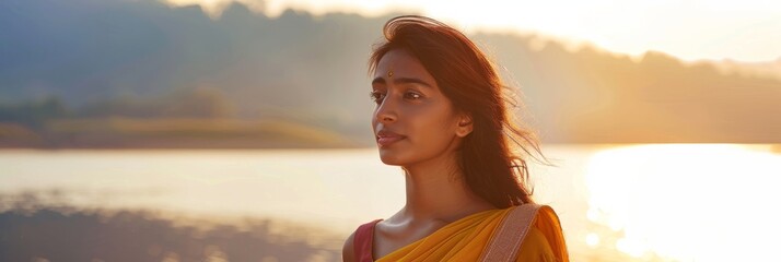 Wall Mural - A young Indian woman stands by a lake at sunset, her serene expression and flowing hair creating a peaceful atmosphere