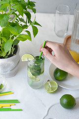 Wall Mural - Hand holding drinking straw, putting in glass with mojito cocktail drink, fresh green limes and mint on home kitchen table background, summer homemade refreshing beverage, lifestyle