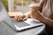 © bongkarn - A cropped image of an Asian woman having coffee while working remotely on her laptop at a cafe.