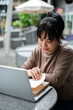 © bongkarn - A woman is working remotely at a cafe, sitting outdoor, using her laptop and reading a book.