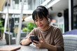 © bongkarn - A positive Asian woman scrolling on her phone while relaxing at an outdoor table in the city.