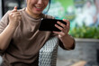 © bongkarn - A cheerful, excited Asian woman is relaxing at an outdoor lounge and watching videos on her phone.