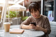 © bongkarn - A positive Asian woman scrolling on her phone while relaxing at an outdoor table in the city.