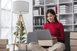 © wichayada - Female Business Owner Managing Online Sales, Sitting on Sofa with Laptop and Packages, Small Business Concept, E-commerce Entrepreneur, Home Office Workspace, Young Woman Running Online Store