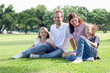 © Stella - Happy family sitting on green grass at the park, smiling father, mother, son and daughter spending time together outdoors in beautiful garden on weekend holiday. Parents and kids have fun outside.