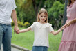 © Stella - Cute little daughter girl holding father and mother hands taking a walk in beautiful summer garden. Joyful kid spends time together with parents at green park. Smiling child with happy family outside.