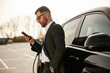 © standret - Leaning on the vehicle while it charges, with smartphone. Businessman in suit is near his black car outdoors