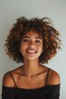 © Creative_Bringer - A young woman with naturally curly hair smiles warmly, expressing happiness and confidence against a plain background