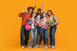 © Prostock-studio - This image shows a group of diverse students smiling and posing together in front of a bright yellow background. They are all holding books and wearing backpacks