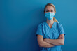 © Alexandra - Female doctor is standing with her arms crossed, wearing a surgical mask and scrubs, in front of a blue background