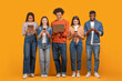 © Prostock-studio - This image shows a group of five young adults standing in a row against a yellow background. They are all looking down at their devices, which include a tablet, a smartphone, and a laptop