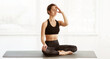 © Prostock-studio - A young woman is sitting cross-legged in a yoga pose on a mat, practicing breathing techniques in a sunlit studio, focused and calm, wearing a black sports outfit, during a serene morning session.