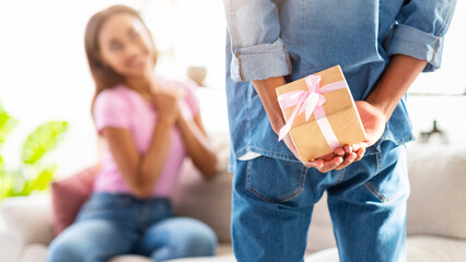 Wall Mural - A guy is hiding a gift behind his back, while a girl sits on the couch in the foreground, looking at him. The girl is out of focus, suggesting a surprise. The setting is a casual living room