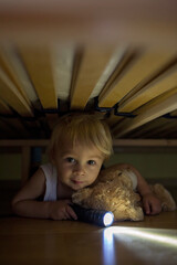 Canvas Print - Little child, hiding under the bed, hugging teddy bear and holding flashlight,