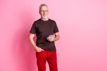 Wall Mural - Portrait of good mood senior man with beard dressed t-shirt in glasses hold smartphone arm in pocket isolated on pink color background