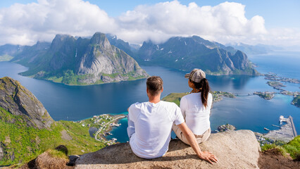 Wall Mural - A couple sits on a mountaintop overlooking a stunning view of the Lofoten Islands in Norway, with the blue ocean and dramatic peaks stretching out in the distance.Reinebringen, Lofoten, Norway